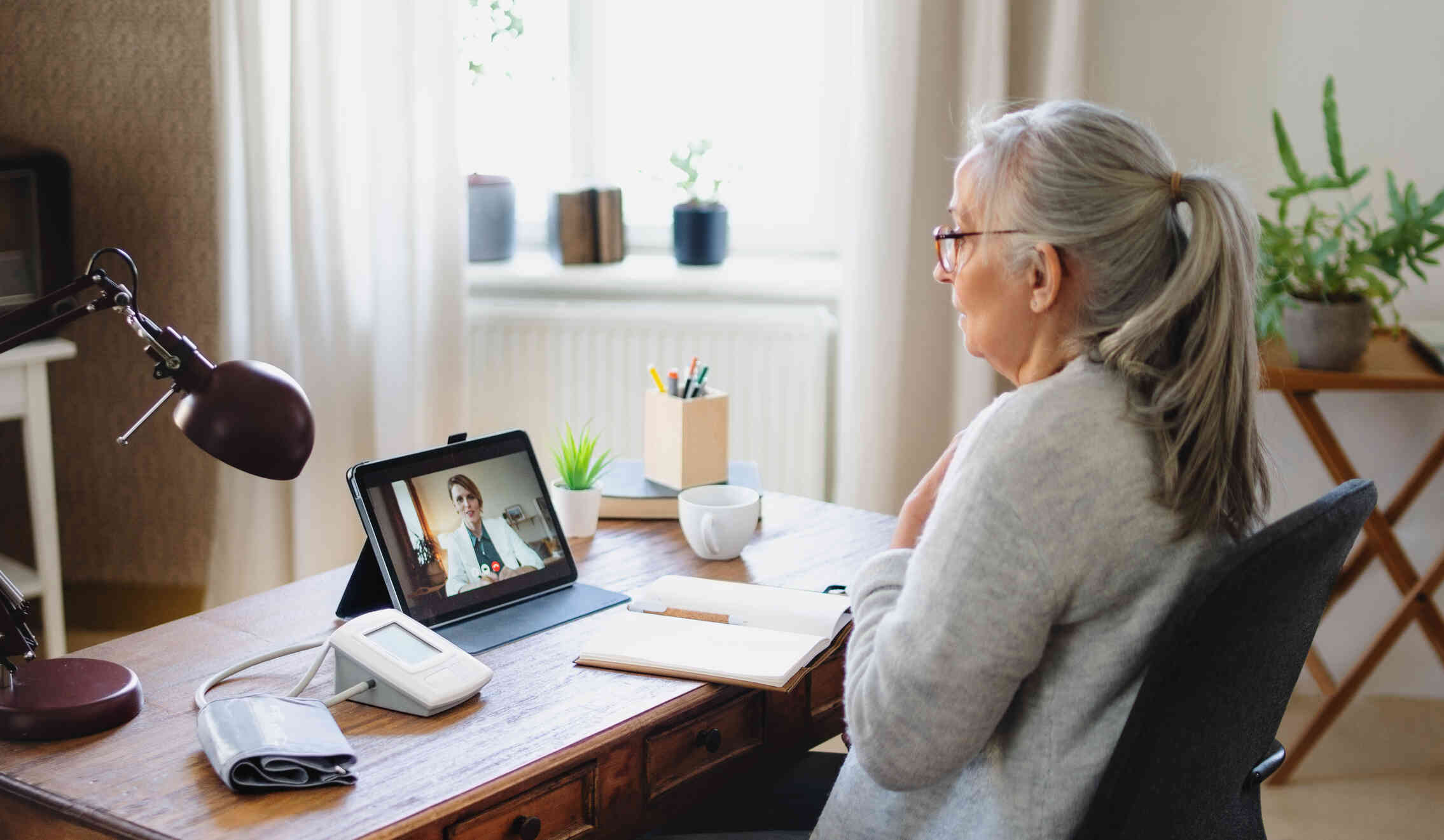 A mature woman sits at a desk with an open notebook and talks to the doctor on the tablet propped up on the table infront of her during a teleheath video call.